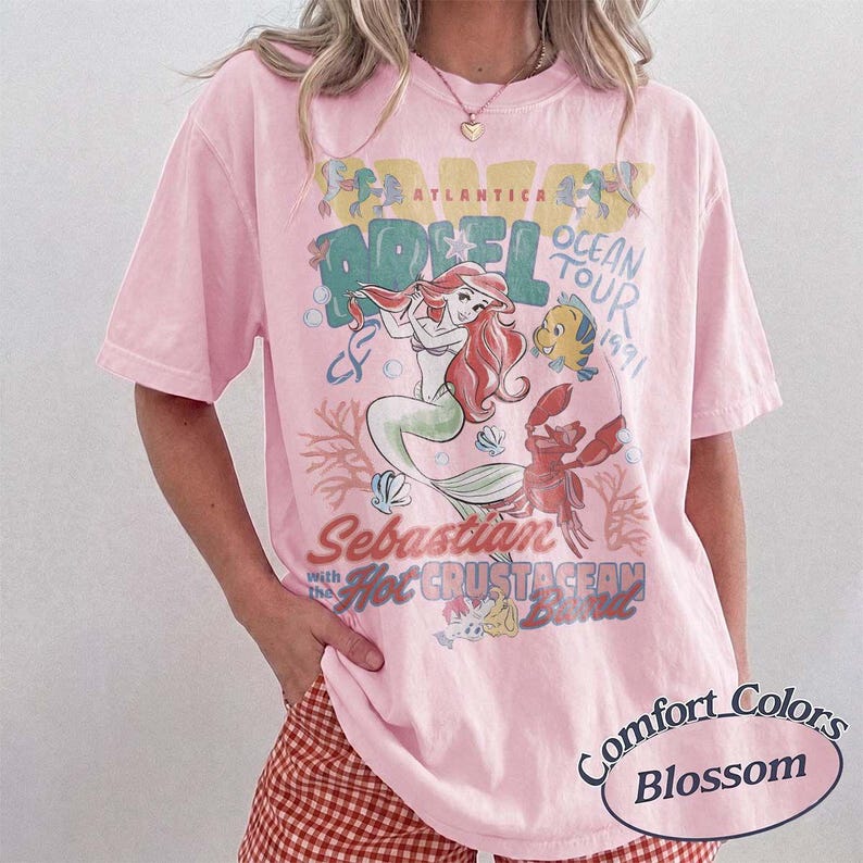 May include: A light pink t-shirt featuring a graphic of Ariel from The Little Mermaid, with the text "Ariel Ocean Tour 1991" and "Sebastian with the Hot Crustacean Band." The shirt is labeled "Comfort Colors Blossom."