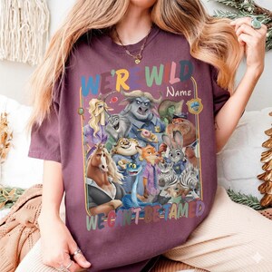May include: A maroon t-shirt featuring a colorful graphic of various cartoon characters. The text "WE'RE WILD" and "WE CAN'T BE TAMED" is displayed above and below the characters. The shirt is made of a soft material.