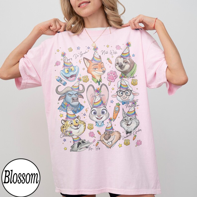 May include: A light pink t-shirt featuring colorful cartoon characters from Zootopia, each wearing a party hat. The shirt includes the text "Gary Di Snake", "Nick Wilde", "Judy Hopps", and "Flash". The Blossom logo is visible.