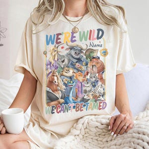 May include: Cream-colored t-shirt featuring a colorful graphic of animated animal characters. The text "WE'RE WILD" and "WE CAN'T BE TAMED" are displayed in rainbow colors. A white mug is held in the person's hand.