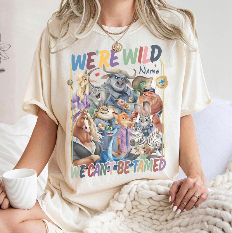 May include: Cream-colored t-shirt featuring a colorful graphic of animated animal characters. The text "WE'RE WILD" and "WE CAN'T BE TAMED" are displayed in rainbow colors. A white mug is held in the person's hand.