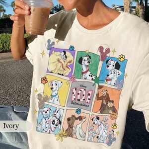 May include: An ivory t-shirt featuring a multi-paneled graphic print of Disney's 101 Dalmatians characters, including Cruella de Vil, various spotted dogs, and the text 101 Dalmatians. The design includes colorful borders and small icons.