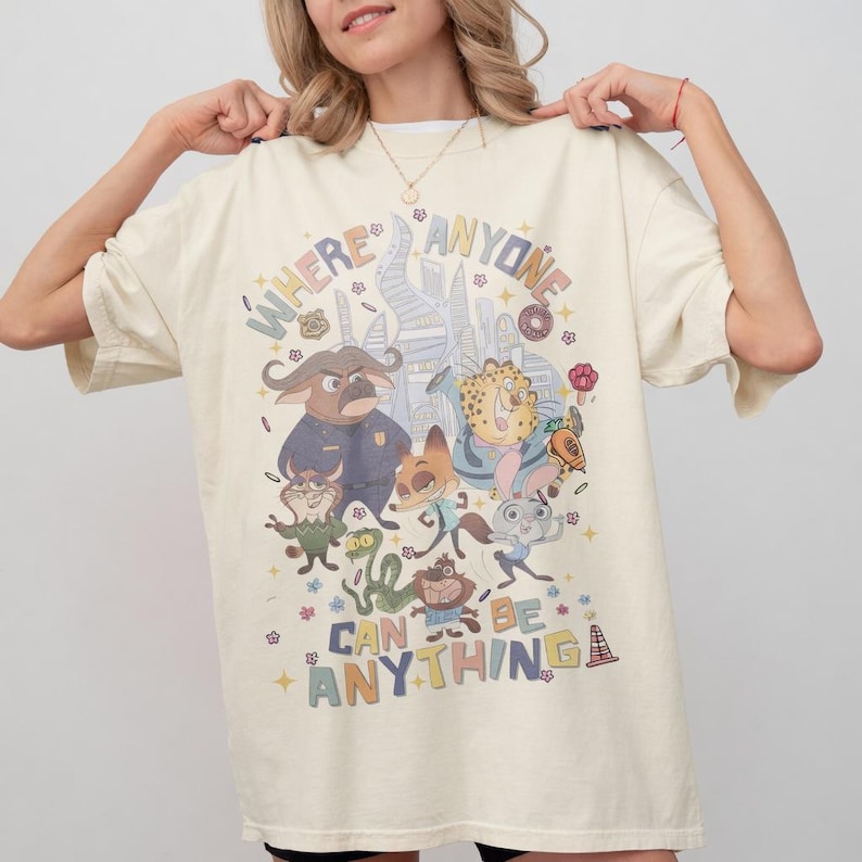 May include: Cream-colored t-shirt featuring a colorful graphic of cartoon characters and the text "WHERE ANYONE CAN BE ANYTHING". The design includes various animal characters and cityscapes, creating a whimsical and playful aesthetic.