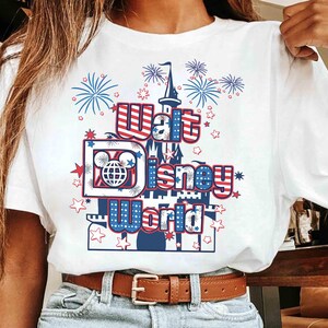 May include: White t-shirt featuring a patriotic design with the text "Walt Disney World." The graphic includes a castle, fireworks, and stars in red, white, and blue. The shirt is worn with blue jeans and a brown belt.
