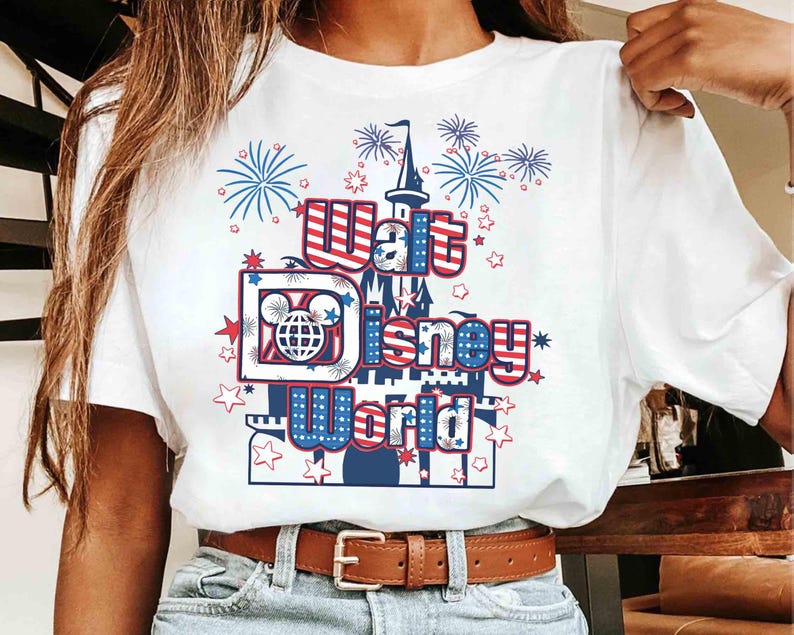 May include: White t-shirt featuring a patriotic design with the text "Walt Disney World." The graphic includes a castle, fireworks, and stars in red, white, and blue. The shirt is worn with blue jeans and a brown belt.