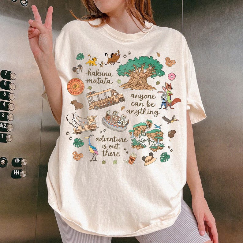 Peut inclure: T-shirt graphique cr&egrave;me avec diverses illustrations de dessins anim&eacute;s et du texte. Le t-shirt comprend les phrases "hakuna matata", "anyone can be anything" et "adventure is out there". Le design incorpore des images d'animaux, d'arbres et de v&eacute;hicules.