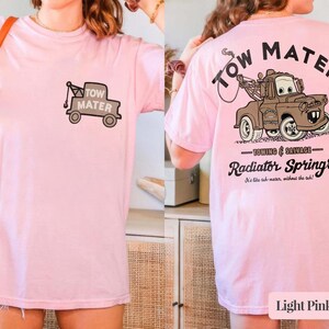 May include: A light pink t-shirt featuring a small graphic of the character Tow Mater on the front left chest. The back displays a large illustration of the brown tow truck with the text Tow Mater, Towing & Salvage, and Radiator Springs.
