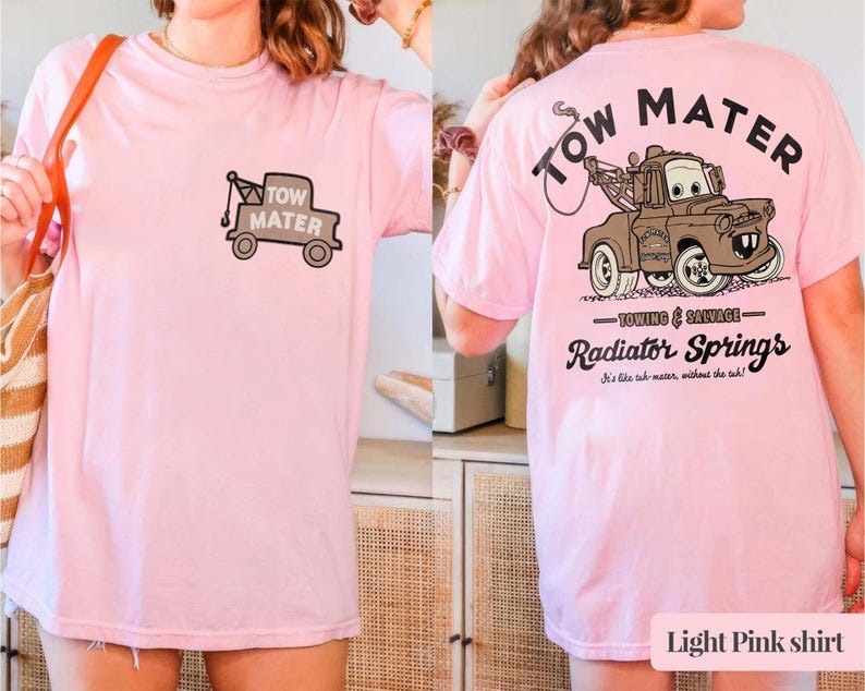 May include: A light pink t-shirt featuring a small graphic of the character Tow Mater on the front left chest. The back displays a large illustration of the brown tow truck with the text Tow Mater, Towing & Salvage, and Radiator Springs.