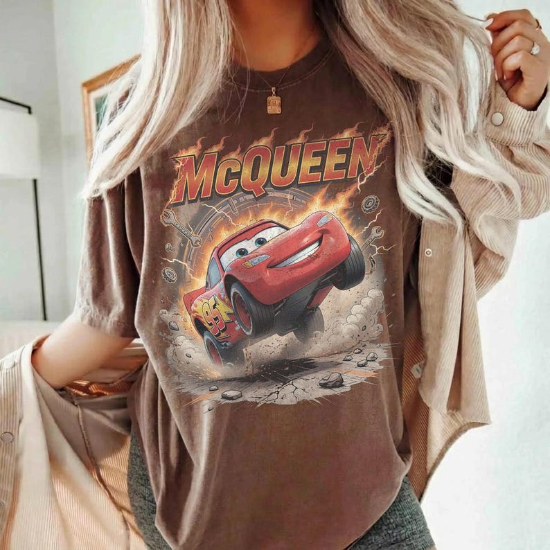 Op de afbeelding: Een bruin t-shirt met korte mouwen en een grafische print van een rode raceauto met het nummer 95 op de zijkant. Het ontwerp bevat vlammen, stofwolken en de tekst MCQUEEN in grote gele en rode letters boven de illustratie.