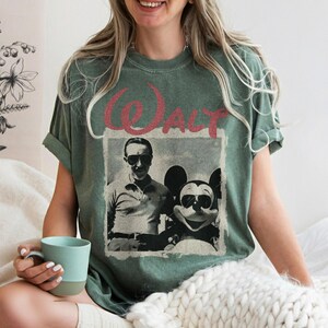 May include: Sage green t-shirt featuring a vintage-style graphic of Walt Disney with Mickey Mouse, both wearing sunglasses. The word "WALT" is printed above the image in a distressed red font. The shirt has rolled-up sleeves.