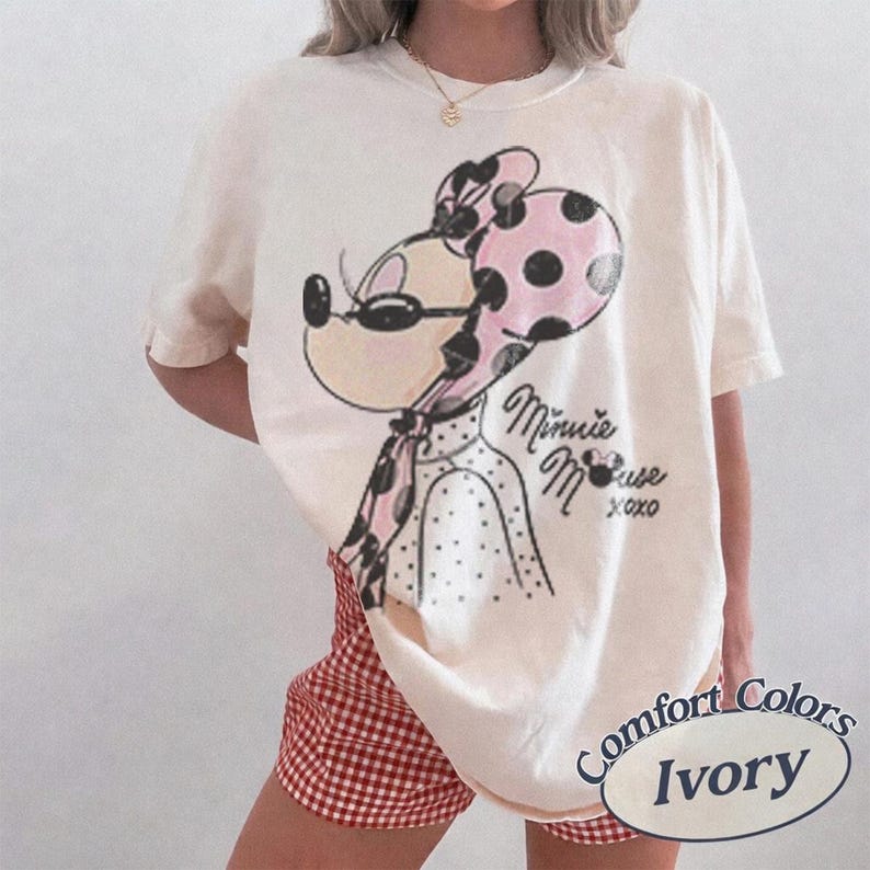 May include: Ivory-colored t-shirt featuring a Minnie Mouse graphic with a pink polka-dot bow and scarf. The text "Minnie Mouse xoxo" is below the graphic. The shirt is paired with red and white checkered shorts.