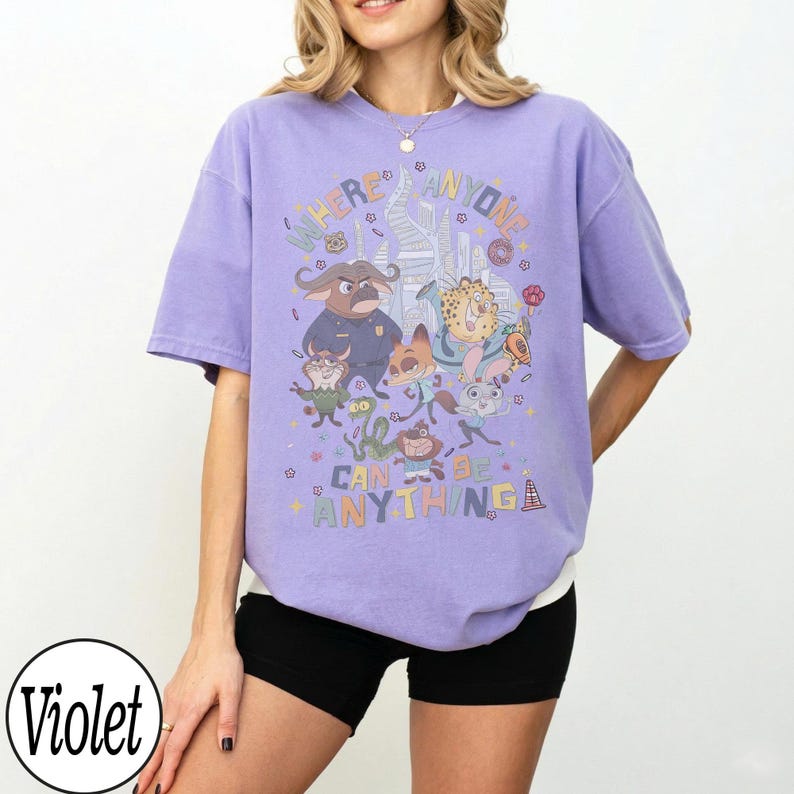 May include: A violet-colored t-shirt featuring a colorful graphic of characters from the animated movie "Zootopia." The graphic includes the text "Where Anyone Can Be Anything." The shirt has short sleeves and a relaxed fit.