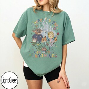 May include: Light green t-shirt featuring a colorful graphic of cartoon characters and the text "WHERE ANYONE CAN BE ANYTHING". The shirt has short sleeves and a relaxed fit, suitable for casual wear.