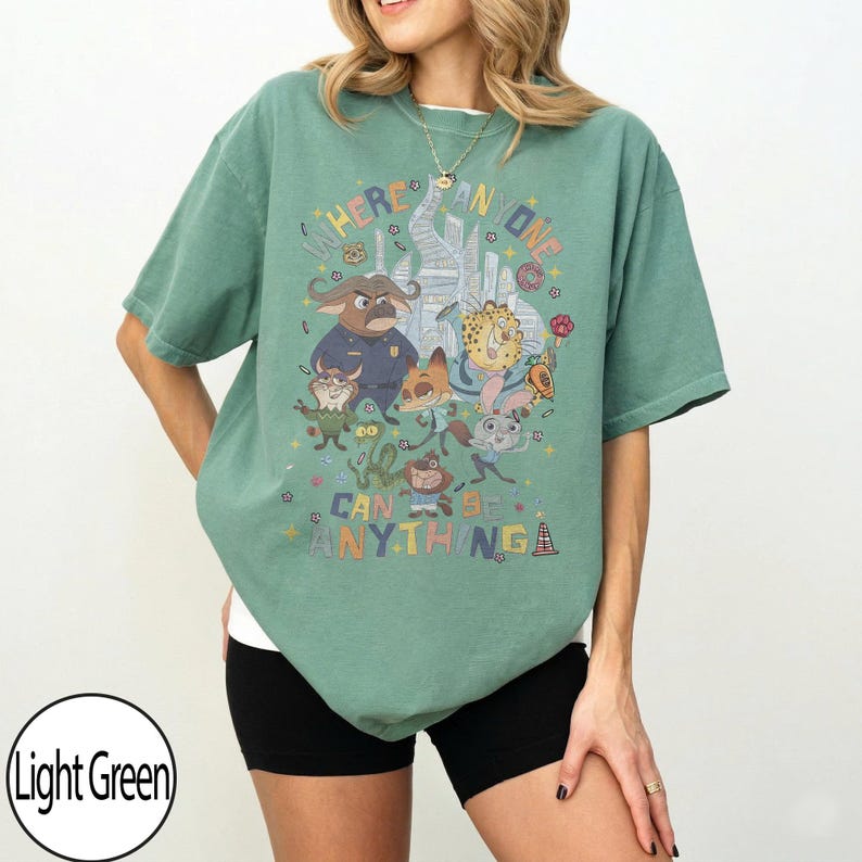May include: Light green t-shirt featuring a colorful graphic of cartoon characters and the text "WHERE ANYONE CAN BE ANYTHING". The shirt has short sleeves and a relaxed fit, suitable for casual wear.