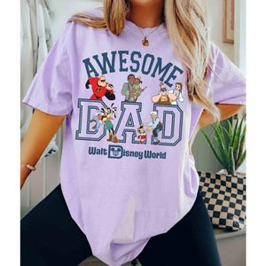 May include: A light purple t-shirt with the words "AWESOME DAD" in a blue and white collegiate style font. The shirt features Disney characters and the text "Walt Disney World" below the word "DAD".