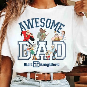 May include: White t-shirt with a blue graphic that reads "AWESOME DAD" with Disney characters and the text "Walt Disney World". The shirt features a brown leather belt and blue jeans.