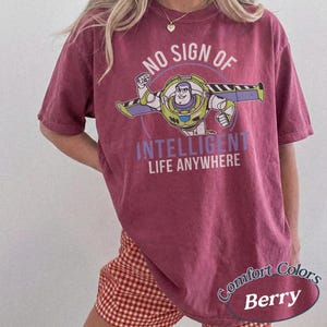 K&ouml;nnte beinhalten: Beerenfarbenes T-Shirt mit einer Grafik von Buzz Lightyear und dem Text "NO SIGN OF INTELLIGENT LIFE ANYWHERE". Das Shirt hat auch die Aufschrift "Comfort Colors" und "Berry". Es ist ein kurz&auml;rmeliges T-Shirt.