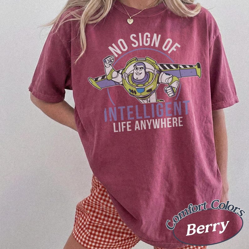 K&ouml;nnte beinhalten: Beerenfarbenes T-Shirt mit einer Grafik von Buzz Lightyear und dem Text "NO SIGN OF INTELLIGENT LIFE ANYWHERE". Das Shirt hat auch die Aufschrift "Comfort Colors" und "Berry". Es ist ein kurz&auml;rmeliges T-Shirt.