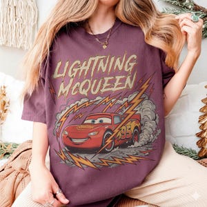May include: A maroon t-shirt featuring a graphic of Lightning McQueen from the Cars movie. The design includes the text "LIGHTNING McQUEEN" above the car, with lightning bolts and smoke effects. The car has the number "95" on its side.