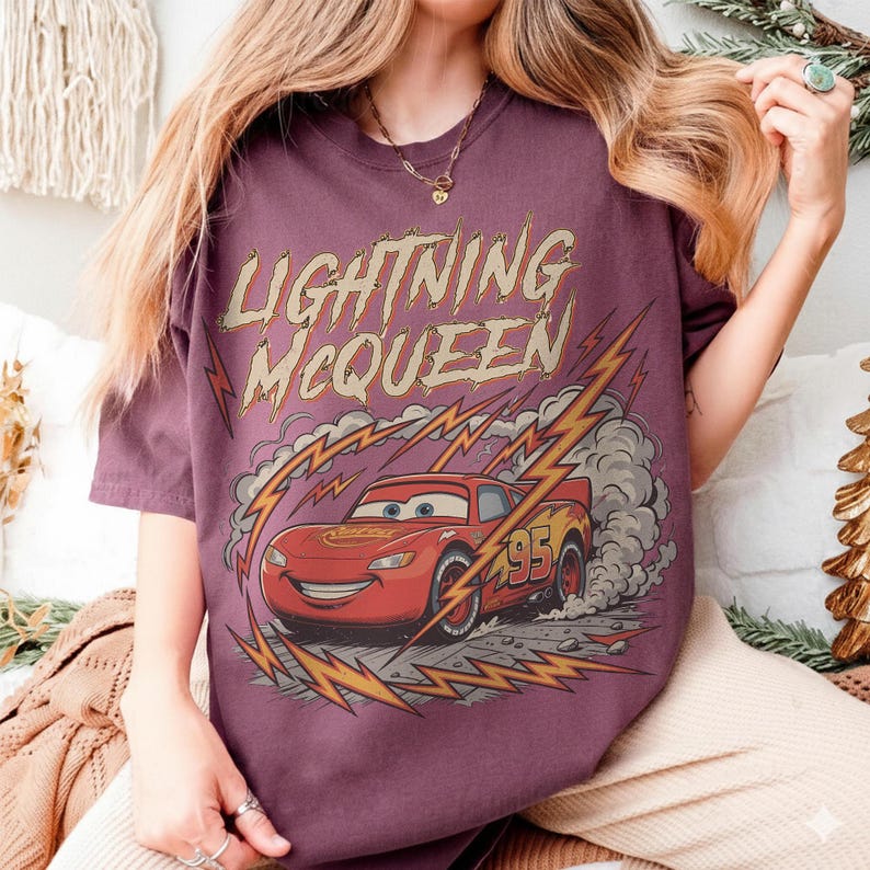 May include: A maroon t-shirt featuring a graphic of Lightning McQueen from the Cars movie. The design includes the text "LIGHTNING McQUEEN" above the car, with lightning bolts and smoke effects. The car has the number "95" on its side.