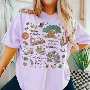 Peut inclure: Un t-shirt violet clair avec des illustrations de dessins anim&eacute;s et du texte. Le t-shirt pr&eacute;sente des images d'animaux, d'un arbre et d'un bus, avec les phrases "hakuna matata" et "adventure is out there".
