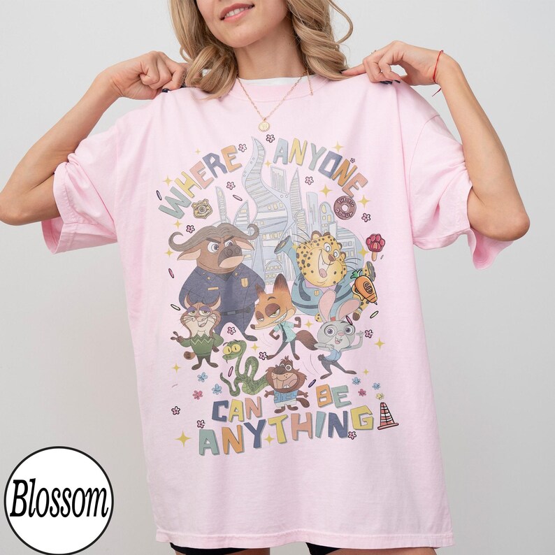 May include: Pink t-shirt featuring a colorful graphic of animated characters and the text "WHERE ANYONE CAN BE ANYTHING". The design includes various animal characters and city buildings. The shirt has short sleeves and a crew neck.