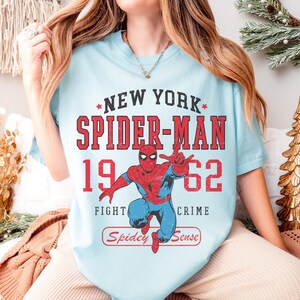 May include: Light blue t-shirt featuring a graphic of Spider-Man in a classic pose. The design includes the text "NEW YORK SPIDER-MAN 1962" in red, along with the phrases "FIGHT CRIME" and "Spidey Sense".