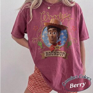 May include: A berry-colored short-sleeve t-shirt featuring a graphic of the character Woody from Toy Story. The design includes a lasso, a sheriff badge, cacti, and the text Woody's Roundup. The shirt has a vintage-style wash and a crew neckline.