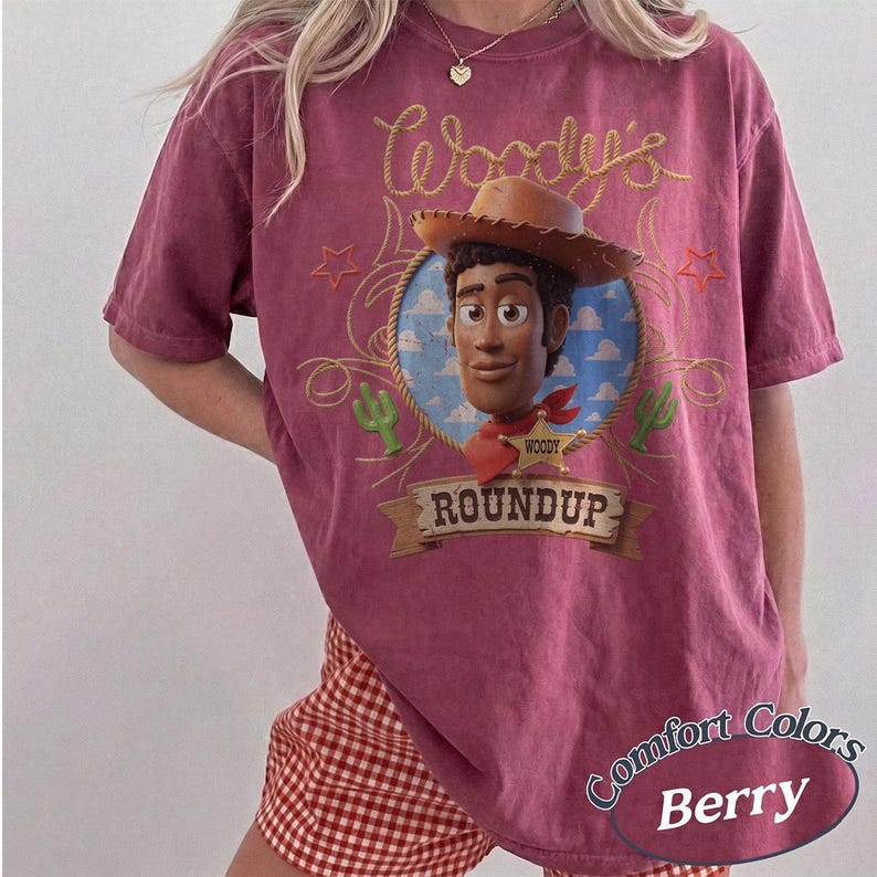 May include: A berry-colored short-sleeve t-shirt featuring a graphic of the character Woody from Toy Story. The design includes a lasso, a sheriff badge, cacti, and the text Woody's Roundup. The shirt has a vintage-style wash and a crew neckline.
