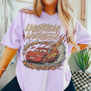 May include: A light purple t-shirt featuring a graphic of Lightning McQueen from the Cars movie. The design includes the text "LIGHTNING McQUEEN" above the car, with lightning bolts and smoke effects.