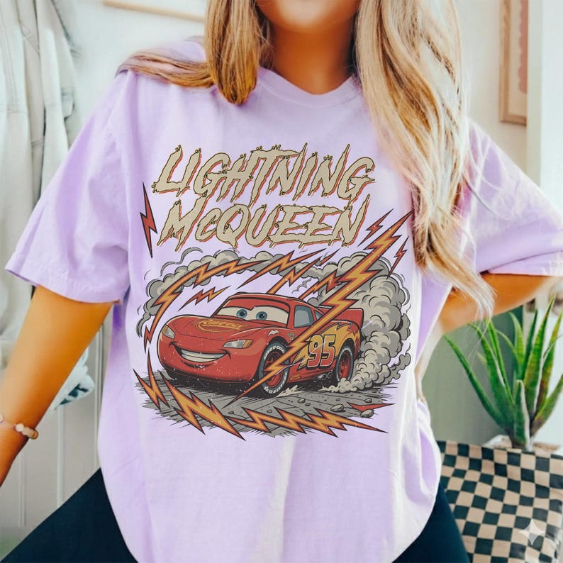 May include: A light purple t-shirt featuring a graphic of Lightning McQueen from the Cars movie. The design includes the text "LIGHTNING McQUEEN" above the car, with lightning bolts and smoke effects.