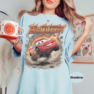 Op de afbeelding: Een lichtblauw t-shirt met een afbeelding van de rode raceauto Lightning McQueen en de tekst MCQUEEN erboven. Het ontwerp bevat vlammen, stofwolken en het nummer 95 op de auto. Het t-shirt wordt gecombineerd met een witte mok met een zon-afbeelding.