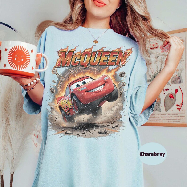 Op de afbeelding: Een lichtblauw t-shirt met een afbeelding van de rode raceauto Lightning McQueen en de tekst MCQUEEN erboven. Het ontwerp bevat vlammen, stofwolken en het nummer 95 op de auto. Het t-shirt wordt gecombineerd met een witte mok met een zon-afbeelding.