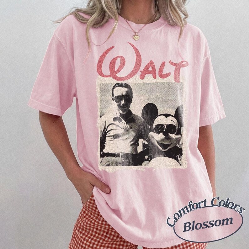 May include: A light pink Comfort Colors t-shirt featuring a vintage black and white image of Walt Disney with Mickey Mouse. The word "Walt" is printed in a distressed red font above the image. The shirt has short sleeves.