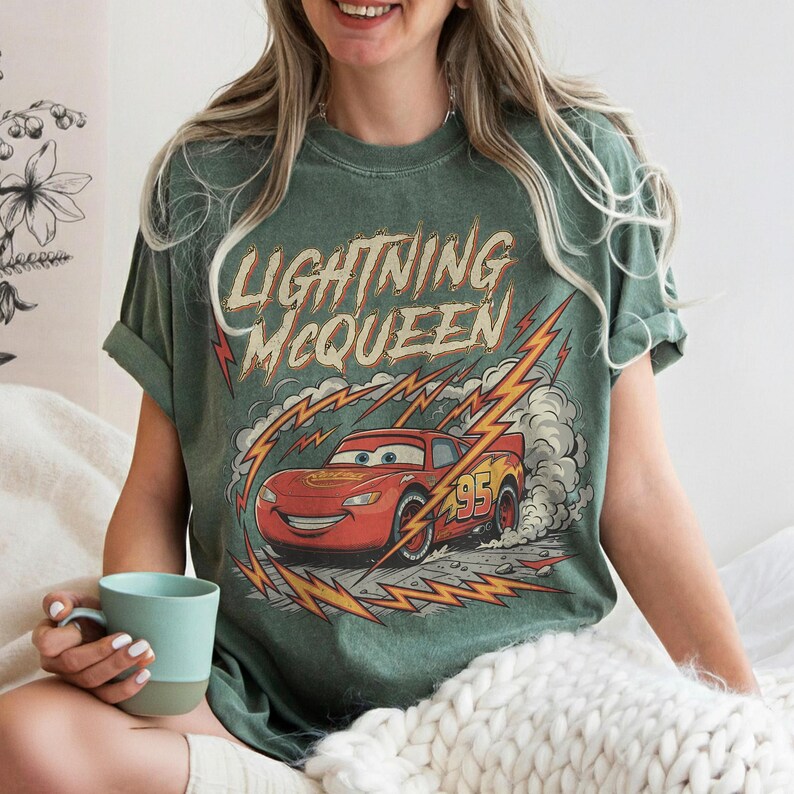 May include: Green t-shirt featuring Lightning McQueen from the Cars movie. The design includes the text "Lightning McQueen" above a red race car with the number 95. The graphic is surrounded by lightning bolts and smoke.