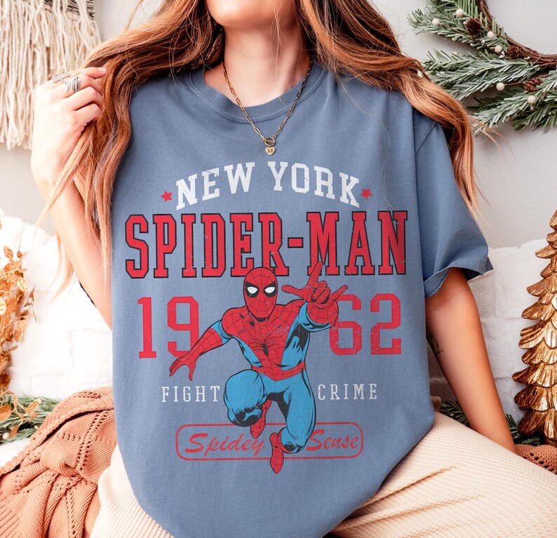 May include: A blue-grey t-shirt featuring a graphic of Spider-Man in a dynamic pose. The design includes the text "NEW YORK SPIDER-MAN 1962" in red and white, with the phrases "FIGHT CRIME" and "Spidey Sense".