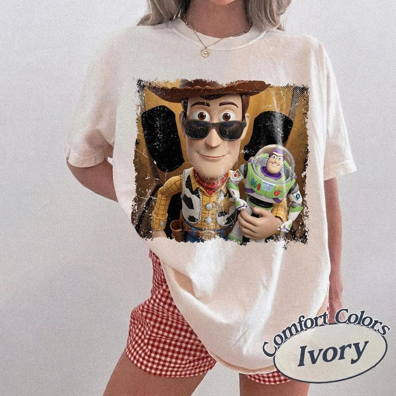 Puede incluir: Camiseta color marfil con un gr&aacute;fico desgastado de Woody y Buzz Lightyear de Toy Story. Woody lleva gafas de sol y un sombrero de vaquero. La camiseta tiene una etiqueta Comfort Colors.