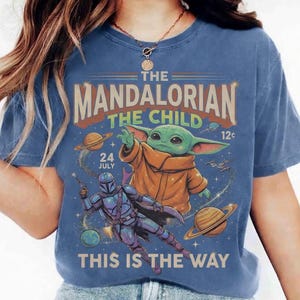 May include: A blue t-shirt featuring a graphic print of The Mandalorian and The Child. The design includes the text THE MANDALORIAN THE CHILD, 24 JULY, 12c, and THIS IS THE WAY, alongside illustrations of planets, stars, and a spaceship in space.