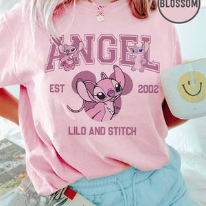 May include: A light pink t-shirt featuring the word "ANGEL" in a purple collegiate font, with images of the character Angel from Lilo & Stitch. The shirt also includes the text "EST 2002" and "LILO AND STITCH".