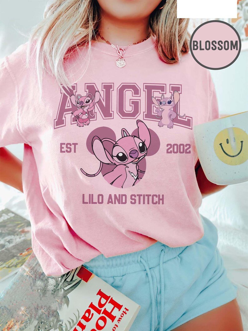 May include: A light pink t-shirt featuring the word "ANGEL" in a purple collegiate font, with images of the character Angel from Lilo & Stitch. The shirt also includes the text "EST 2002" and "LILO AND STITCH".