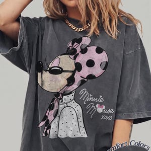 May include: Dark gray t-shirt featuring a Minnie Mouse graphic with a pink polka dot bow and scarf. The shirt has the words "Minnie Mouse xoxo" in a script font. The shirt is labeled "Comfort Colors Pepper".