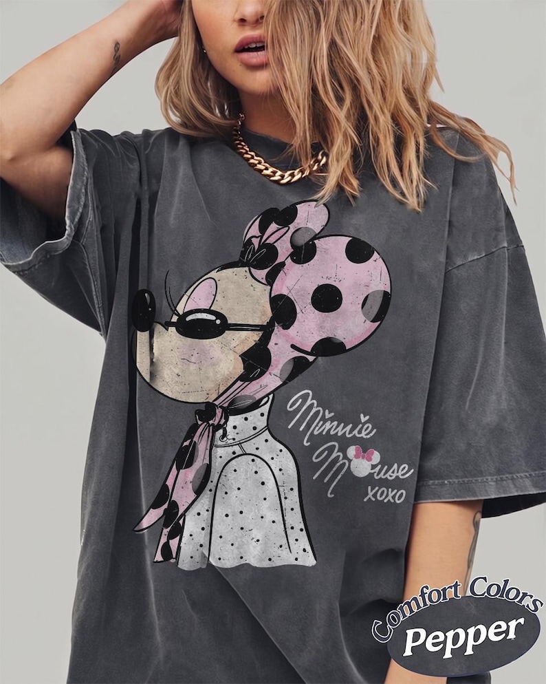 May include: Dark gray t-shirt featuring a Minnie Mouse graphic with a pink polka dot bow and scarf. The shirt has the words "Minnie Mouse xoxo" in a script font. The shirt is labeled "Comfort Colors Pepper".