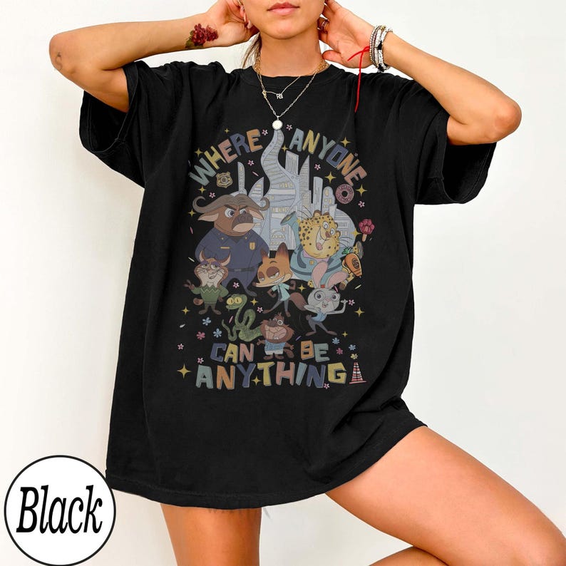 May include: Black t-shirt featuring a colorful graphic with the text "WHERE ANYONE CAN BE ANYTHING." The design includes cartoon characters and a cityscape. The shirt is a comfortable, oversized fit.