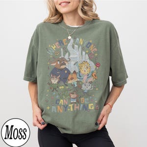 May include: Olive green t-shirt featuring a colorful graphic of cartoon characters and the text "WHERE ANYONE CAN BE ANYTHING". The design includes a cityscape and various animal figures. The shirt has short sleeves and a relaxed fit.