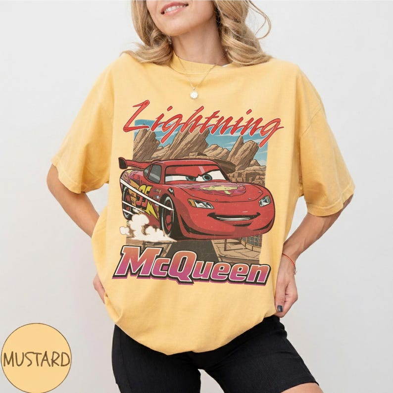 K&ouml;nnte beinhalten: Senfgelbes T-Shirt mit einer Grafik von Lightning McQueen aus dem Film Cars. Das Design enth&auml;lt den Text "Lightning McQueen" in Rot und "McQueen" in Lila. Das T-Shirt hat einen Vintage-Look.