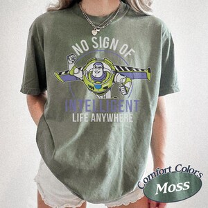 K&ouml;nnte beinhalten: Ein moosgr&uuml;nes T-Shirt mit einem Buzz Lightyear-Motiv und dem Text "NO SIGN OF INTELLIGENT LIFE ANYWHERE". Das Design zeigt eine kreisf&ouml;rmige Grafik von Buzz Lightyear. Das Shirt ist mit "Comfort Colors Moss" gekennzeichnet.