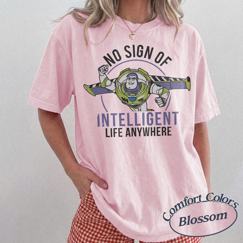 K&ouml;nnte beinhalten: Hellrosa Comfort Colors Blossom T-Shirt mit einem Buzz Lightyear-Motiv und dem Text "NO SIGN OF INTELLIGENT LIFE ANYWHERE". Das Design zeigt einen kreisf&ouml;rmigen Rahmen um die Figur.
