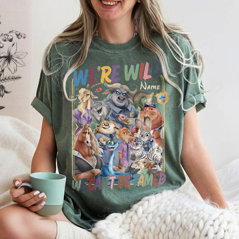 May include: Green t-shirt with a colorful graphic featuring cartoon animal characters and the text "WE'RE WILD" and "W CAN'T BE TAMED". The shirt has a crew neck and short sleeves.