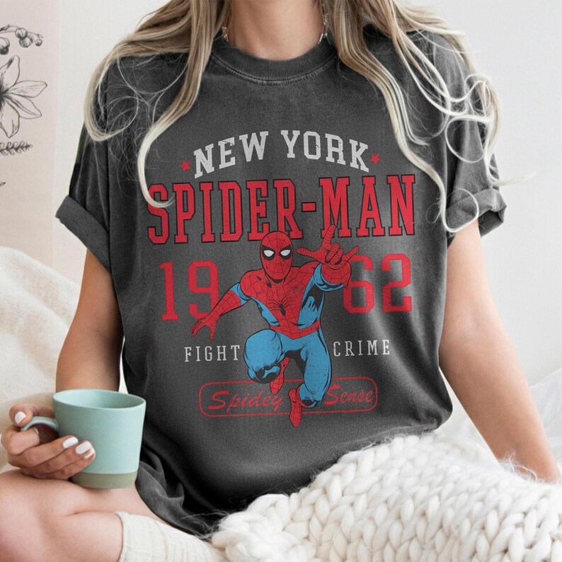 May include: Dark gray t-shirt featuring a graphic of Spider-Man in a dynamic pose. The design includes the text "NEW YORK SPIDER-MAN 1962 FIGHT CRIME" in red and white, with "Spidey Sense" below the character.