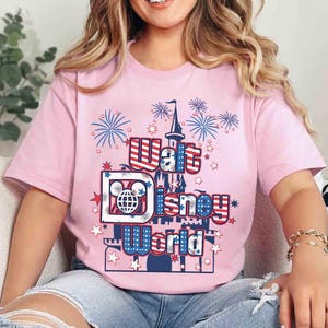 May include: A light pink t-shirt featuring the text "Walt Disney World" in red, white, and blue, with a castle and fireworks graphic. The shirt has short sleeves and is made of a soft material.